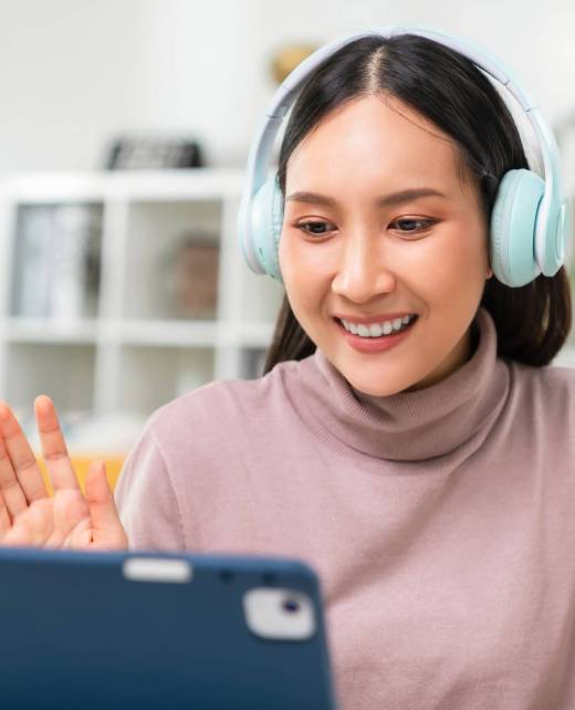 Cheerful beautiful Asian woman wearing headphones and look to digital tablet at video calling meeting and study online on the Internet.